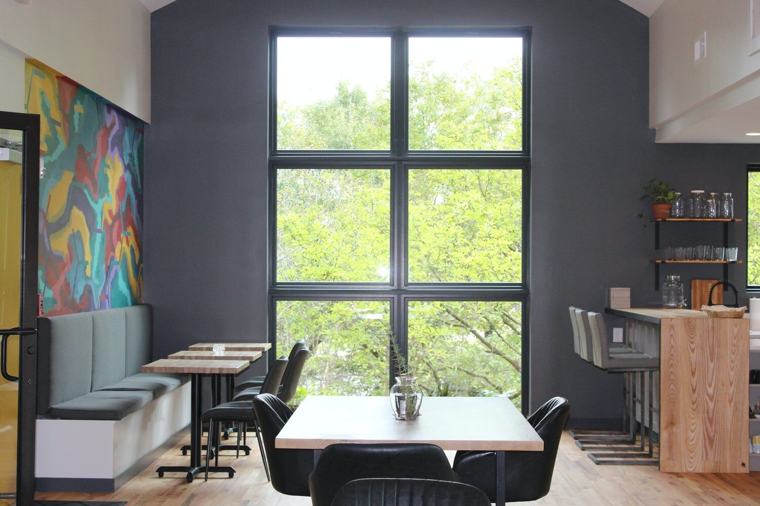 Tables by tall windows, with a colorful mural on the wall
