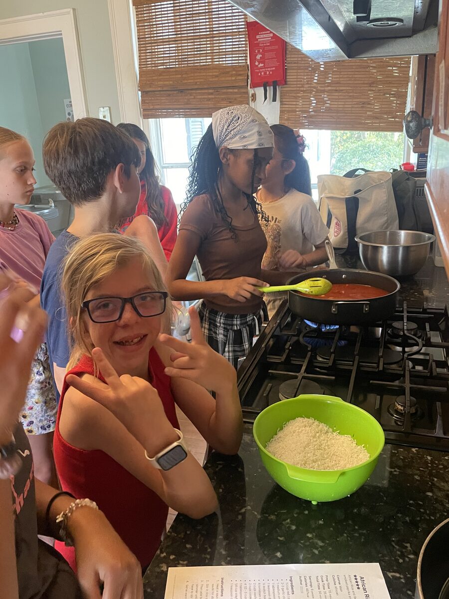 Kids crowded round the stove cooking a pot of sauce, one grinning at the camera