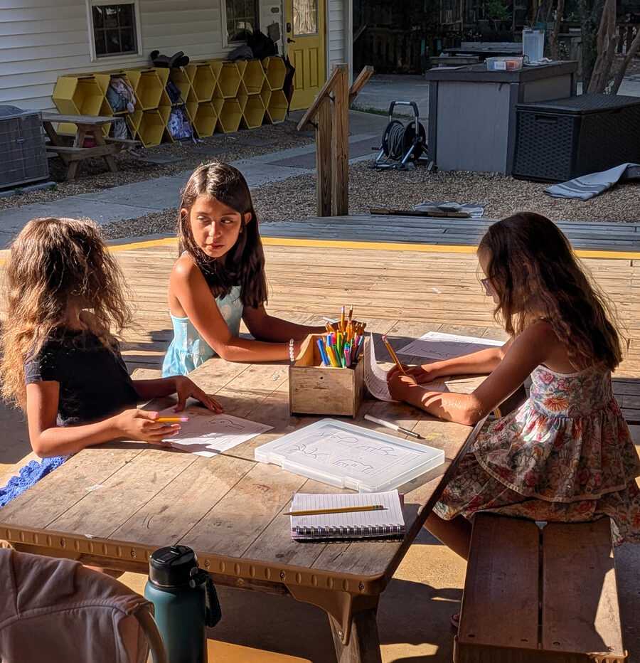 Three kids drawing together at a picnic table in the shade