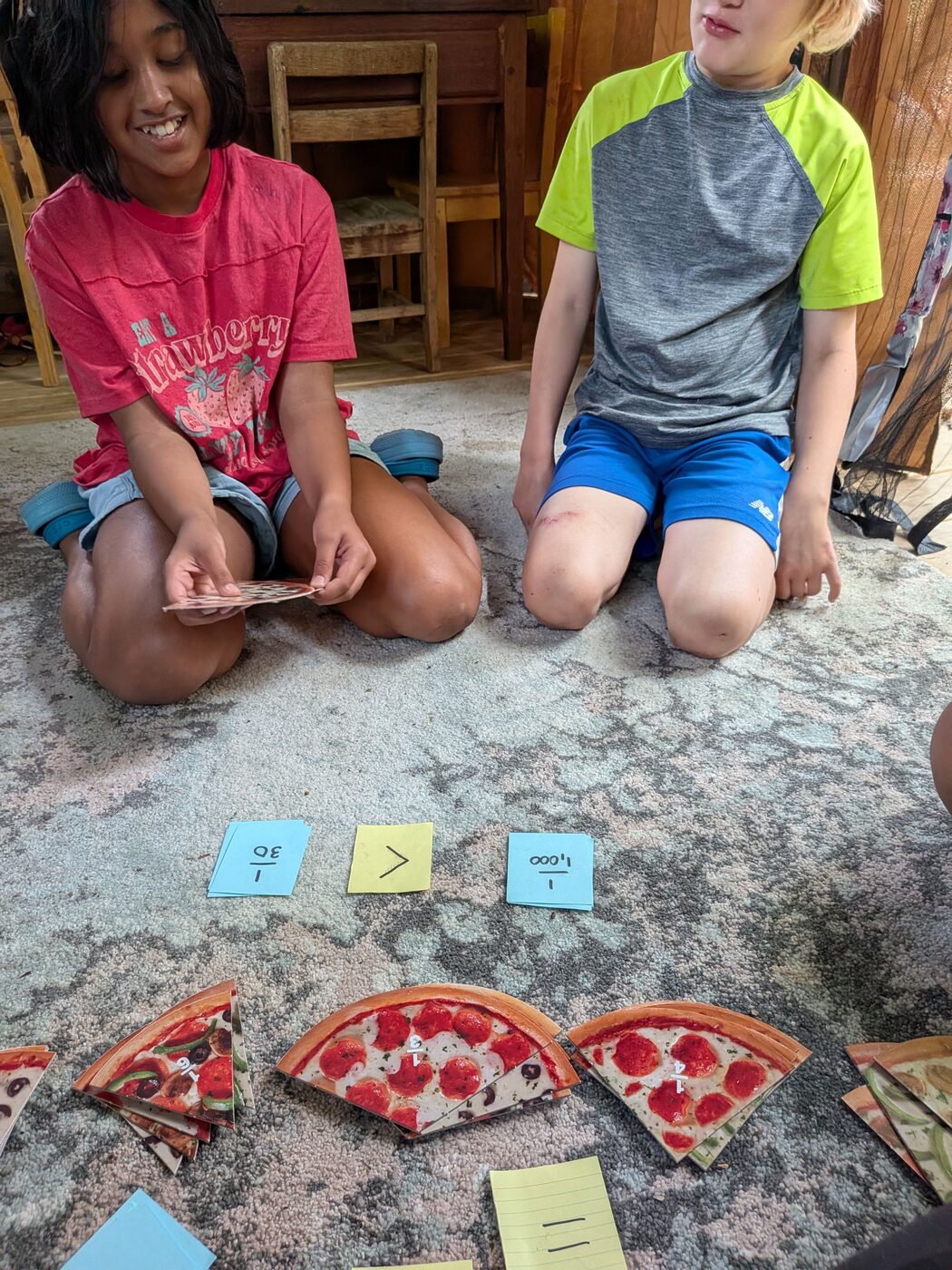 Two kids comparing fractions with paper pizza slices on the rug